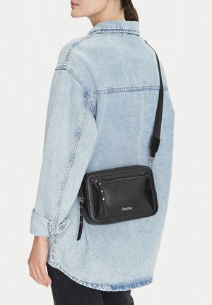 Mujer con una chaqueta ligera de mezclilla y un bolso cruzado negro DeeZee colgado de su hombro, sobre un fondo liso.