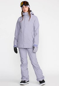 Ski suit in light lavender fabric features a fitted jacket with a hood, matching pants, and gloves. Accessories include goggles and a beanie.