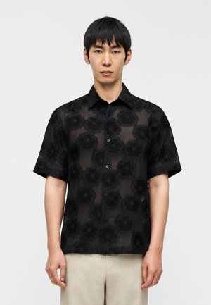 Μαύρο πουκάμισο με κοντά μανίκια και λουλουδάτο σχέδιο, φτιαγμένο από διάφανο ύφασμα, με μπροστινό κούμπωμα με κουμπιά και κλασικό γιακά.