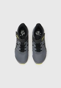 Zapatillas deportivas grises con acentos negros, parte superior de malla, punta redonda y suela plana. Cuenta con un cuello acolchado y una correa de gancho y bucle. Detalle amarillo en la suela.