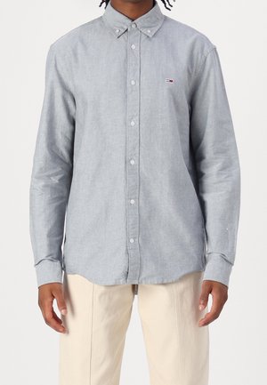 Mann trägt ein hellblaues Oxford-Hemd mit Knöpfen und einem kleinen rot-weiß-blauen Logo auf der Brust, kombiniert mit beigefarbenen Hosen.