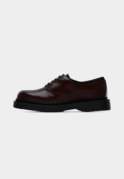 Brauner polierter Leder-Oxford-Schuh mit schwarzer Sohle und Schnürsenkeln, Seitenansicht vor schlichter hellgrauer Hintergrund.