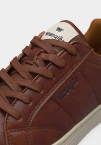 Καφετί δερμάτινο αθλητικό παπούτσι με υφασμένα καφέ κορδόνια, λογότυπο Wrangler στο πλάι και στη γλώσσα, και λευκή σόλα που φαίνεται σε λευκό φόντο.