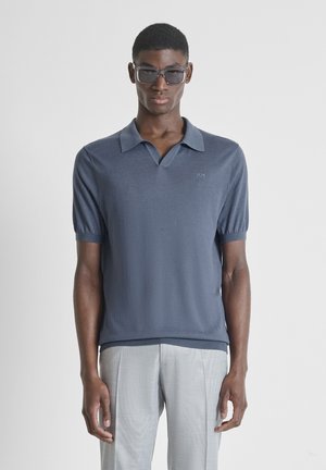 Joven con gafas de sol oscuras, camiseta polo azul de manga corta y pantalones gris claro, de pie contra un fondo liso.