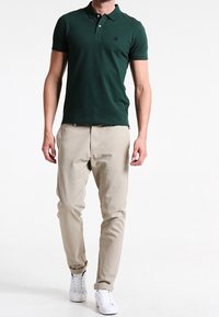 Polo shirt vert foncé avec un col à deux boutons, assorti à un pantalon chino beige clair et des baskets blanches. Coupe décontractée, matière en mélange de coton.