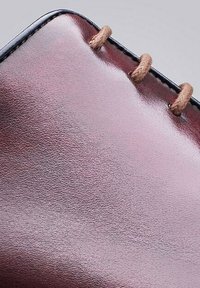Leren product met een glanzende bordeauxrode afwerking, voorzien van bruine leren veters die door de rand zijn geregen en de gestructureerde oppervlakte accentueren.