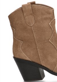 Bota de ante marrón hasta el tobillo con un diseño de costura en zigzag y un tacón en bloque negro y curvado. La bota cuenta con una lengüeta para facilitar su puesta.