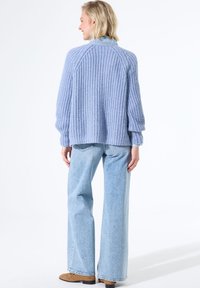 Femme de dos portant un pull en laine épaisse bleu clair, un jean large délavé et des chaussures brunes sur fond blanc.