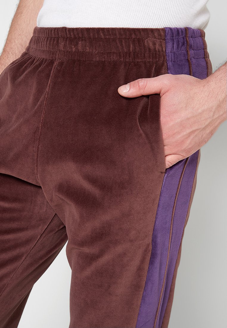 Pantalons de jogging en velours marron avec une bande latérale violette, taille élastique et poches latérales, avec une texture douce et lisse.