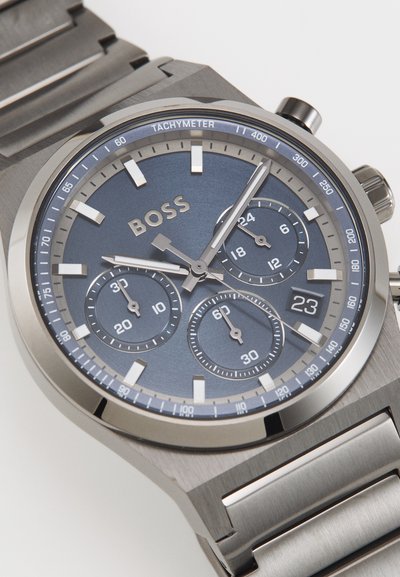 Montre chronographe métallique avec un cadran bleu, des accents en argent, trois sous-cadrans et un bracelet en acier inoxydable à texture brossée. Marque présentée : BOSS.