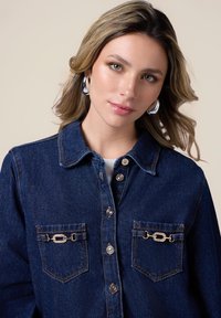 Jachetă din denim de culoare albastru închis cu guler, închidere cu butoane argintii și două buzunare frontale cu accente decorative de lanț.