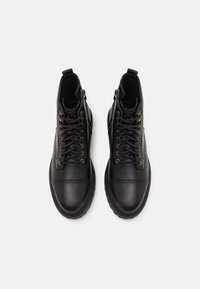 Botas negras de cuero hasta el tobillo con puntera redondeada, cordones en la parte delantera, ojales en tono dorado y detalle de cremallera lateral. Textura suave con costuras mínimas.