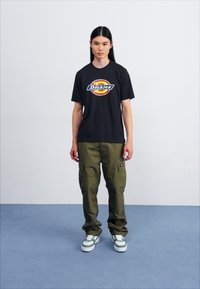 Svart t-skjorte med korte ermer og en fargerik "Dickies" logo, kombinert med grønne cargo-bukser og hvite joggesko med grønne detaljer.