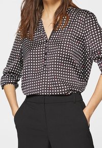 Kvinna klädd i en svartvit rutmönstrad blus med uppvikta ärmar, innanför högmidjade svarta byxor, med händerna i fickorna.