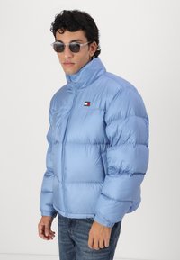 Helle blaue Steppjacke mit hohem Kragen, gestepptem Textil und kleinem gesticktem Logo. Kombiniert mit dunklen Jeans und Sonnenbrille.