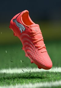 Ehelekirju roosa jalgpallikets, millel on tekstureeritud pind, nööbitav disain ja must Puma logo, hõljumas rohu kohal lendavate mullaosakestega.