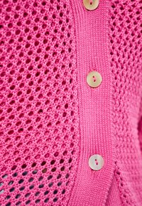 Różowy sweter robiony na drutach, z koronkowym wzorem ażurowym, z solidnymi ściągaczami oraz okrągłymi, jasnymi guzikami z przodu.