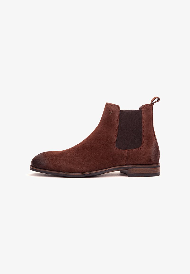 Bota Chelsea de ante marrón con paneles laterales elásticos negros y lengüeta para tirar, tacón bajo de madera y punta redondeada sobre un fondo blanco.