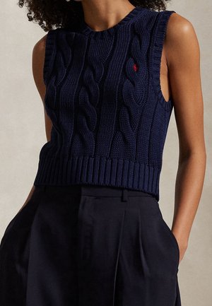 Gilet sans manches en maille torsadée bleu marine porté avec un pantalon noir taille haute, mains dans les poches, fond neutre.