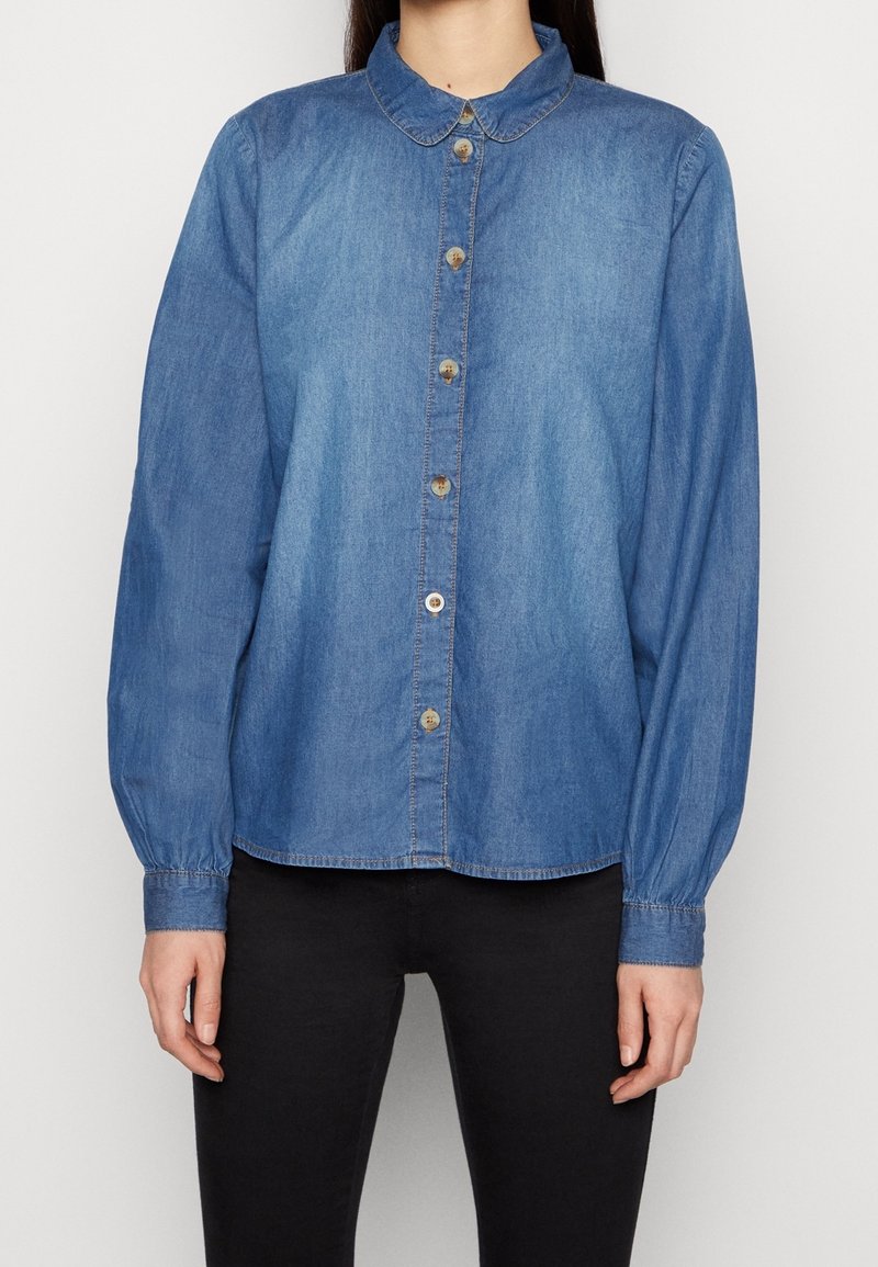 Camicia in denim azzurro chiaro con collo a puntale, maniche lunghe, chiusura frontale a bottoni e polsini elastici, caratterizzata da bottoni marroni e un orlo leggermente curvo.