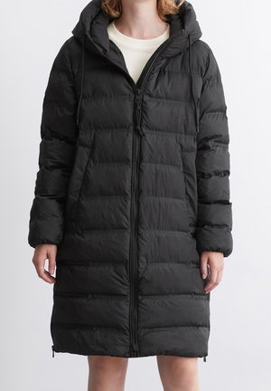 Personne portant un long manteau matelassé noir avec capuche et fermeture éclair à l'avant, par-dessus un haut blanc côtelé. Mains relâchées sur les côtés.