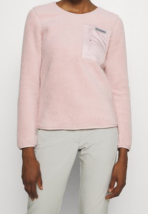 Osoba nosząca jasnoróżową, puszystą bluzkę z długim rękawem z jasnoróżową kieszonką na zamek błyskawiczny na lewej piersi oraz jasnoszare spodnie.