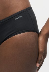 Κοντινό πλάνο ενός προσώπου που φοράει μαύρα εσώρουχα Organic Basics, δείχνοντας το πλαϊνό ύψος του ισχίου και το χέρι που στηρίζεται απαλά στο πάνω μέρος του μηρού.