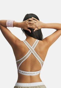 Athletic woman with long dark hair stands with hands clasped behind head, wearing crisscross sports bra and pink wristband.