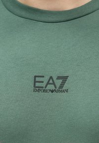 Zöld pamut póló bordázott, kerek nyakkivágással. A mellrészen fekete EA7 Emporio Armani logóval, sima textúrával és testhezálló fazonnal.