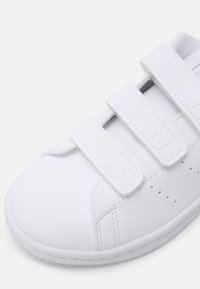 Λευκά συνθετικά αθλητικά παπούτσια με τρεις ιμάντες Velcro, στρογγυλή μύτη, λεία επιφάνεια και διάτρητες λεπτομέρειες στα πλάγια.