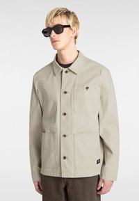 Beige canvas jacket with a collar, front button closure, and two large pockets. Black tag on the lower side. Worn with dark shades.