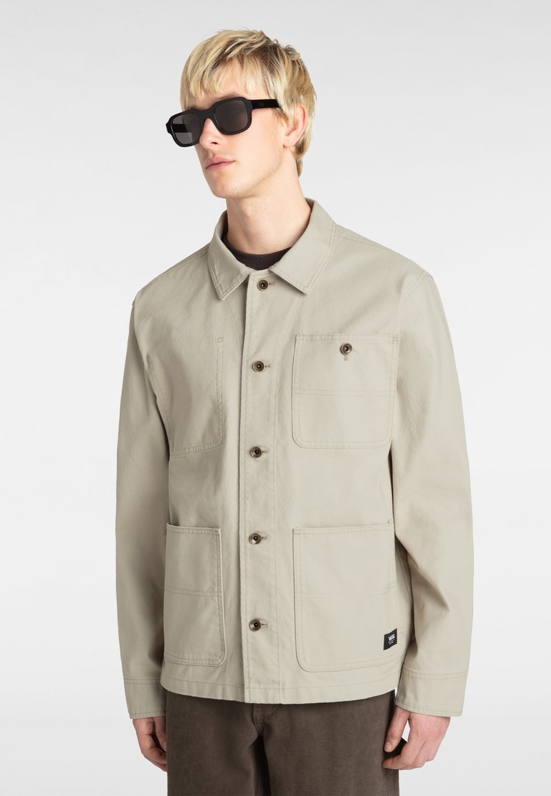 Beige canvas jacket with a collar, front button closure, and two large pockets. Black tag on the lower side. Worn with dark shades.