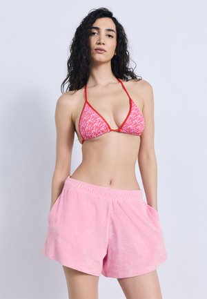 Mujer con cabello largo, rizado y oscuro, vistiendo un top de bikini con estampado rojo y rosa y pantalones cortos de tela tipo toalla rosa, de pie con las manos en los bolsillos.