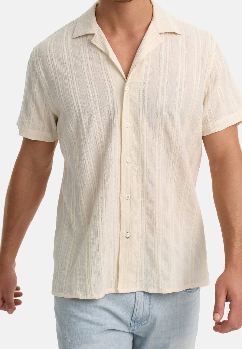 Homme portant une chemise crème à manches courtes, boutonnée, avec une texture subtile à rayures verticales et un col, associée à un jean bleu clair.