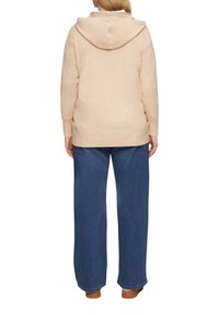 Beige Hoodie mit einer großen Kapuze, seitlichen Schlitzen und einem lockeren Schnitt, kombiniert mit blauen weiten Jeans und braunen Slippern.