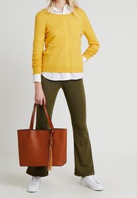 Geel sweater, witte overhemdkraag, groene gebloemde uitlopende broek en een bruine tas met franjes, aangevuld met witte sneakers.