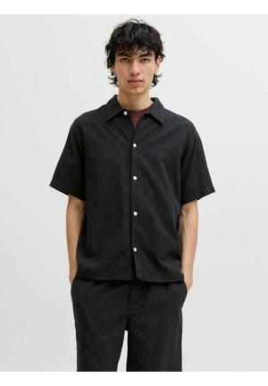 Ung mand med mørkt krøllet hår iført en kortærmet sort skjorte med knapper og matchende sorte bukser, stående mod en ensfarvet lys baggrund.