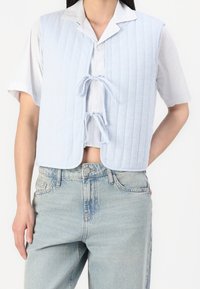 Kobieta nosząca jasnoniebieski pikowany bezrękawnik związany z przodu na białej koszuli z krótkim rękawem zapinanej na guziki oraz jasnych dżinsach.