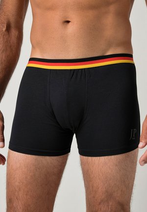 Hombre que lleva calzoncillos bóxer negros con cintura de rayas rojas, negras y amarillas, y logo JP 1880 en el muslo izquierdo.