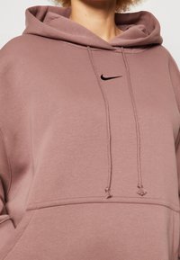Rosa Kapuzenpullover aus strukturiertem Stoff, mit einer Kängurutasche und einem kleinen schwarzen Nike-Logo auf der Brust. Mit Kordelzügen ausgestattet.