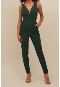 Kvinna klädd i en ärmlös, mörkgrön jumpsuit med djup V-halsringning och fickor, i kombination med guld höga klackar och minimalistisk smyckesdesign.