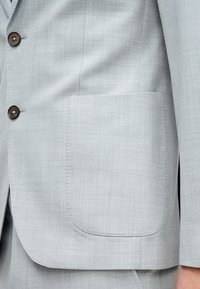 Világosszürke öltönyzakó texturált anyagból, két barna gombbal és bal oldalon egy négyzet alakú zsebbel. Tiszta, modern dizájn.
