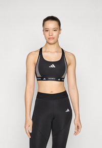 Black sports bra with grey accents, ADIDAS logo, and adjustable straps; paired with high-waisted black leggings featuring textured waistband.