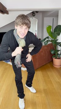 Cămașă neagră cu mâneci lungi, pantaloni negri și adidași albi. Un fular cu model este așezat în jurul gâtului. Într-un spațiu interior cu o plantă și o canapea.