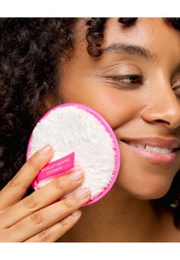 Disco redondo y suave para la limpieza facial con una textura blanca y esponjosa y un borde exterior rosa, sostenido contra la mejilla de una persona. Etiqueta de la marca visible.