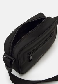Borsa a tracolla nera con tessuto testurizzato, scomparti con zip, una tracolla staccabile e un logo discreto sul lato.