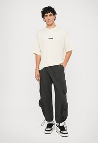 T-shirt beige a maniche corte con testo nero "BARROW", abbinato a pantaloni cargo neri con tasche applicate e polsini con coulisse. Sneakers in bianco e nero.