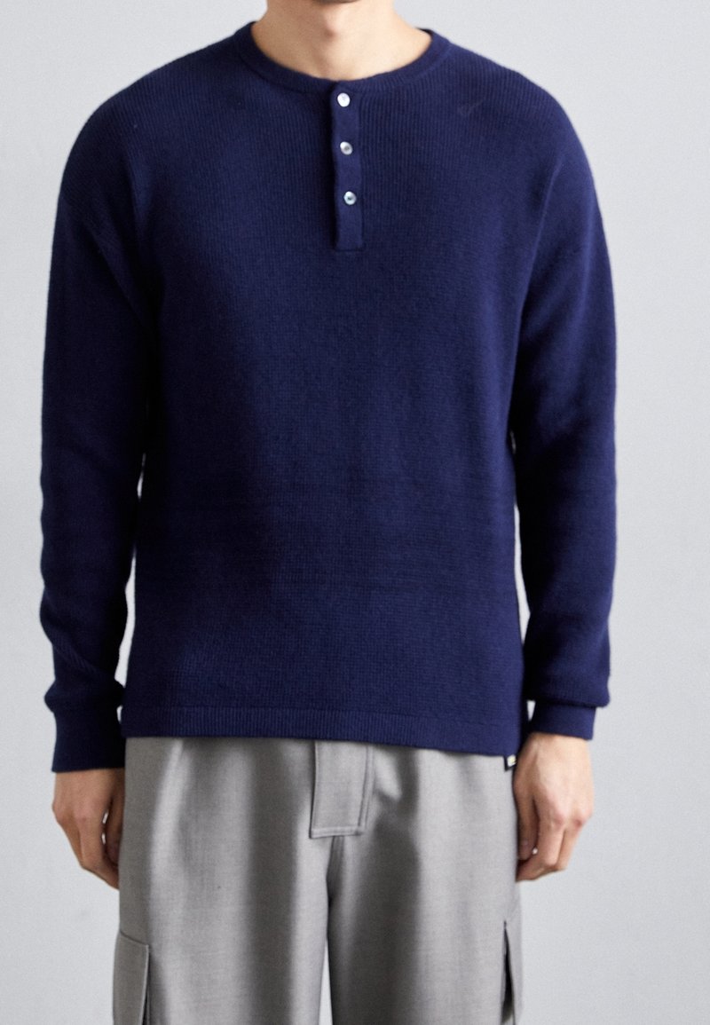 Homme portant un henley à manches longues bleu marine avec trois boutons et un pantalon gris clair sur un fond uni.