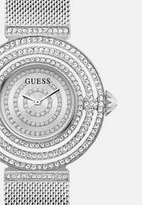 Srebrny zegarek z siatki z okrągłymi wzorami, ozdobiony cyrkoniami, z prostą tarczą i czarnymi wskazówkami pokazującymi czas. Nazwa marki: GUESS.