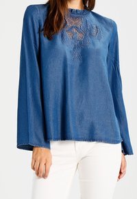 Blouse en jean bleu à manches longues, ornée d'une broderie florale délicate sur la poitrine et d'un col à volants. Texture lisse, coupe ample.